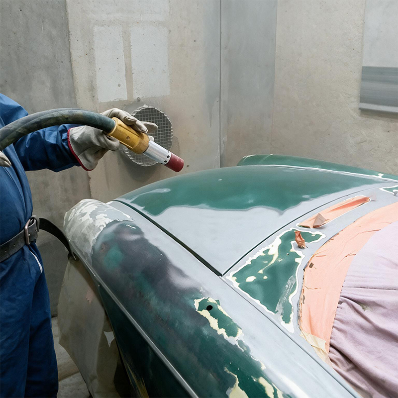 Technicien réalisant l’aérogommage de la carrosserie verte d’une voiture ancienne avant restauration